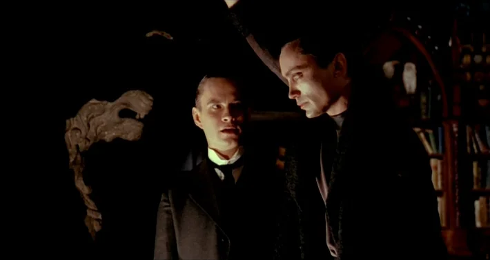 Blood For Dracula (1974) movie still: medium — A dark study features a bookshelf in the background and a carved wooden statue. Two men stand in the…