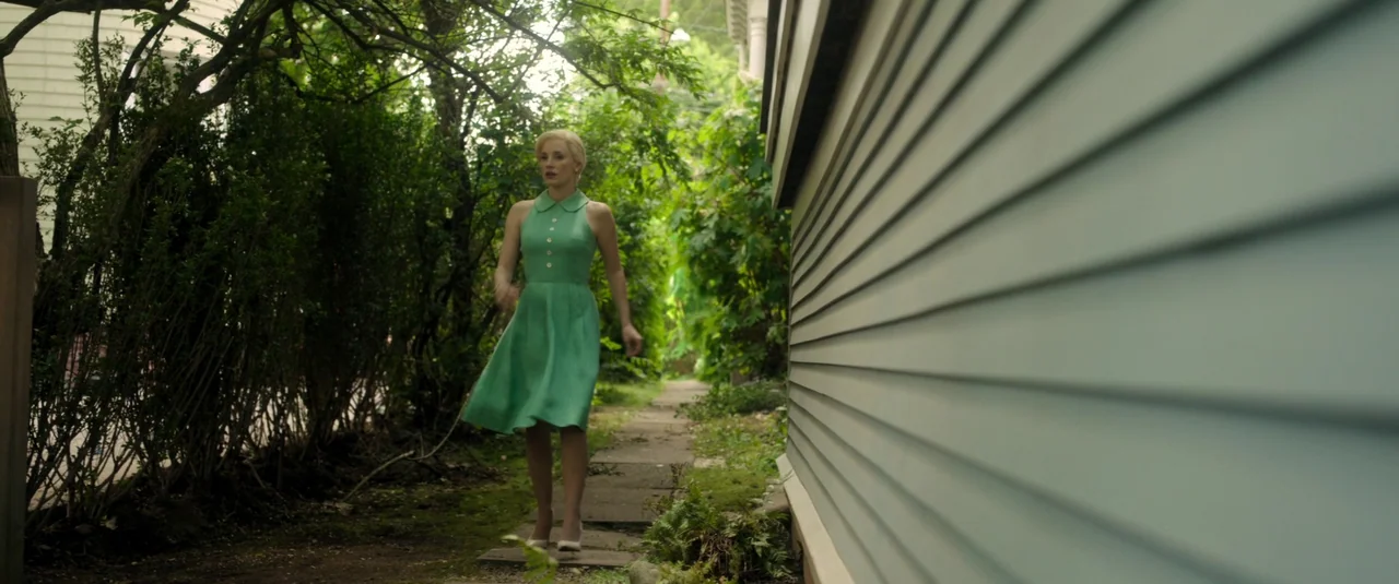 Mother’s Instinct (2024) movie still: fullbody — A narrow alleyway runs between a dense row of trees and the exterior wall. A woman in a green dress…