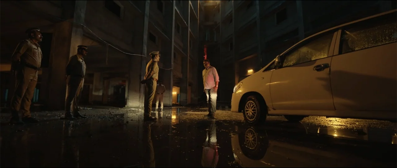 Garudan (2024) movie still: wide — An unfinished concrete parking structure at night with water pooling on the ground. Three police officers in…