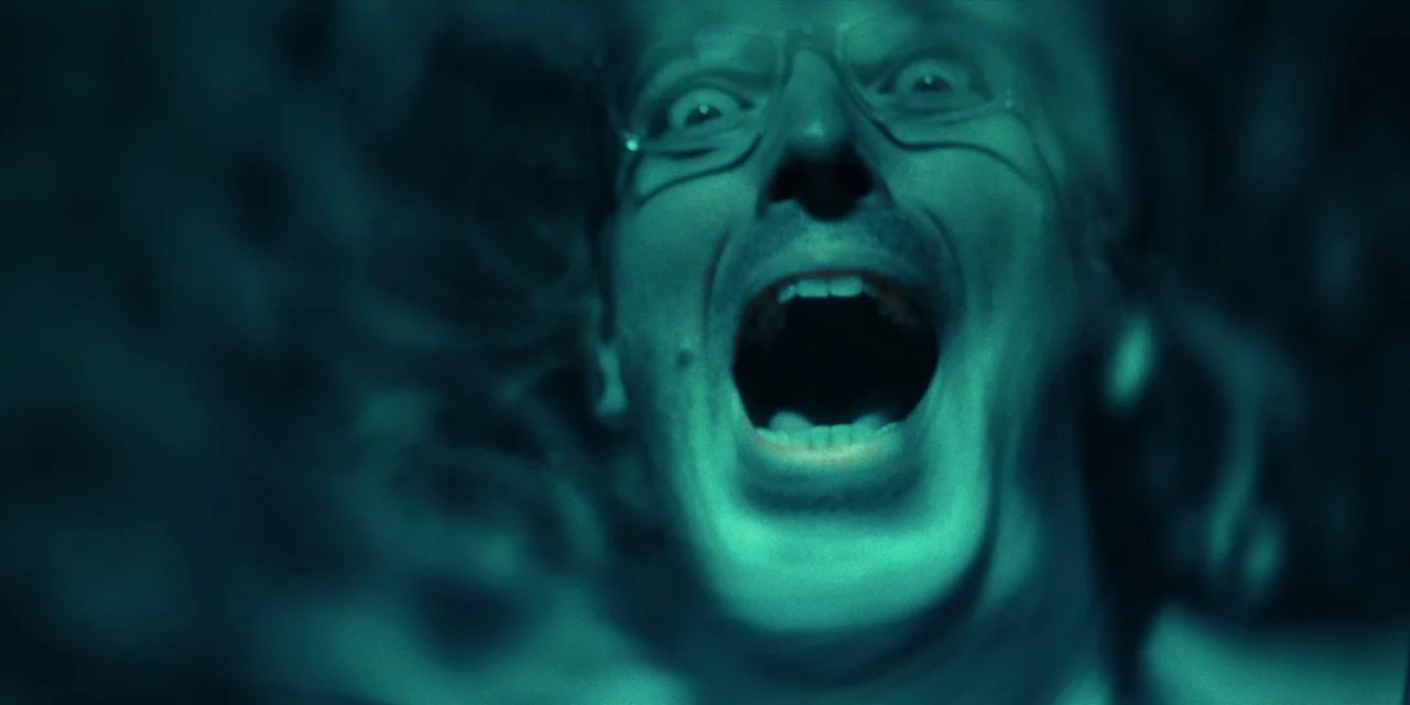 Destroy All Neighbours (2024) movie still: closeup — A dark, nondescript interior space is dominated by a single, brightly lit face. A man with…