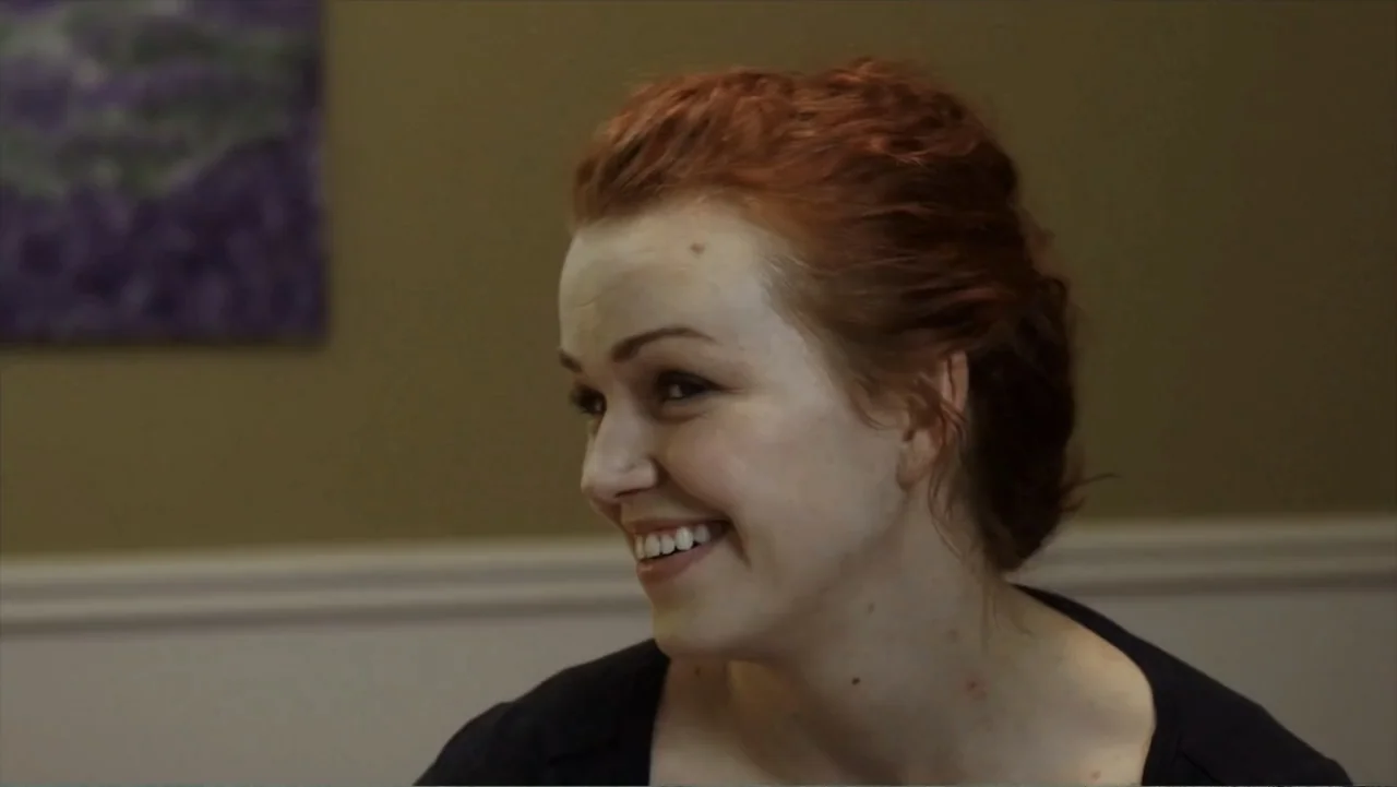 Hello I Love You (2018) movie still: closeup — A plain interior room with a beige wall featuring a blurry framed artwork. A woman with red hair…