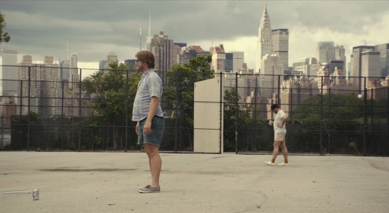 The Comedy (2012) movie still: wide — A paved handball court stands against a background of urban apartment buildings and skyscrapers. One man…