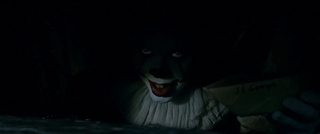 It (2017) movie still: closeup — A dark, cramped storm drain space is swallowed by heavy shadows. A clown figure emerges from the darkness, offering…