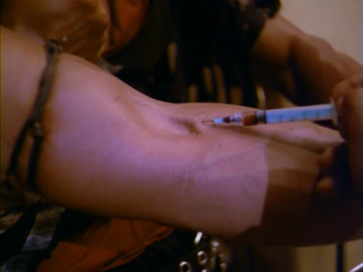 Tenement (1985) movie still: closeup — A close view of a person's muscular arm inside a nondescript room. A hypodermic needle penetrates the skin of…