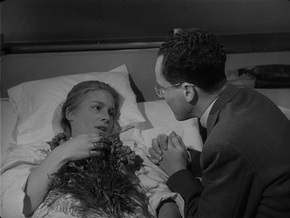 Brink of Life (1958) movie still: medium — A sterile medical room features a white pillow and bed linens. A woman lies in bed holding a bouquet of…