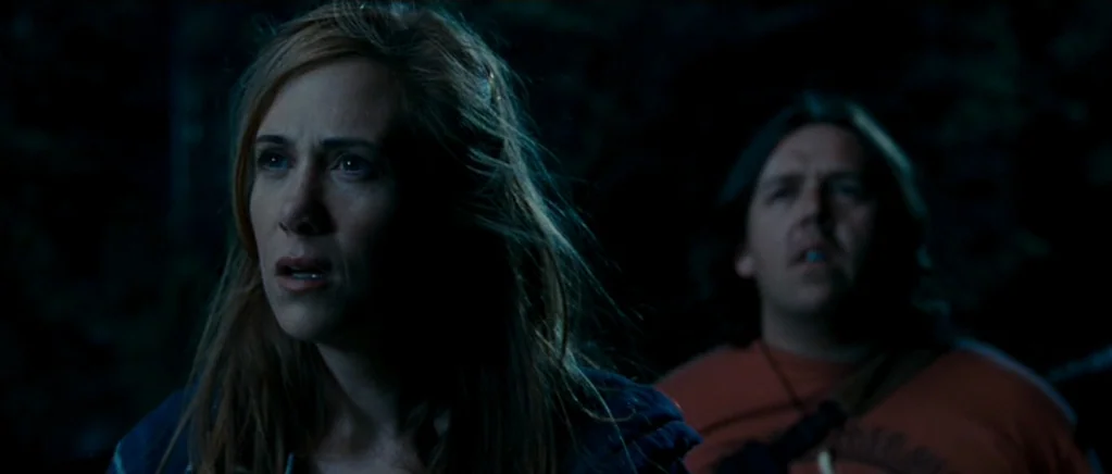 Paul (2011) movie still: medium — A dark, wooded environment at night with blurred foliage in the background. A woman and a man look toward…