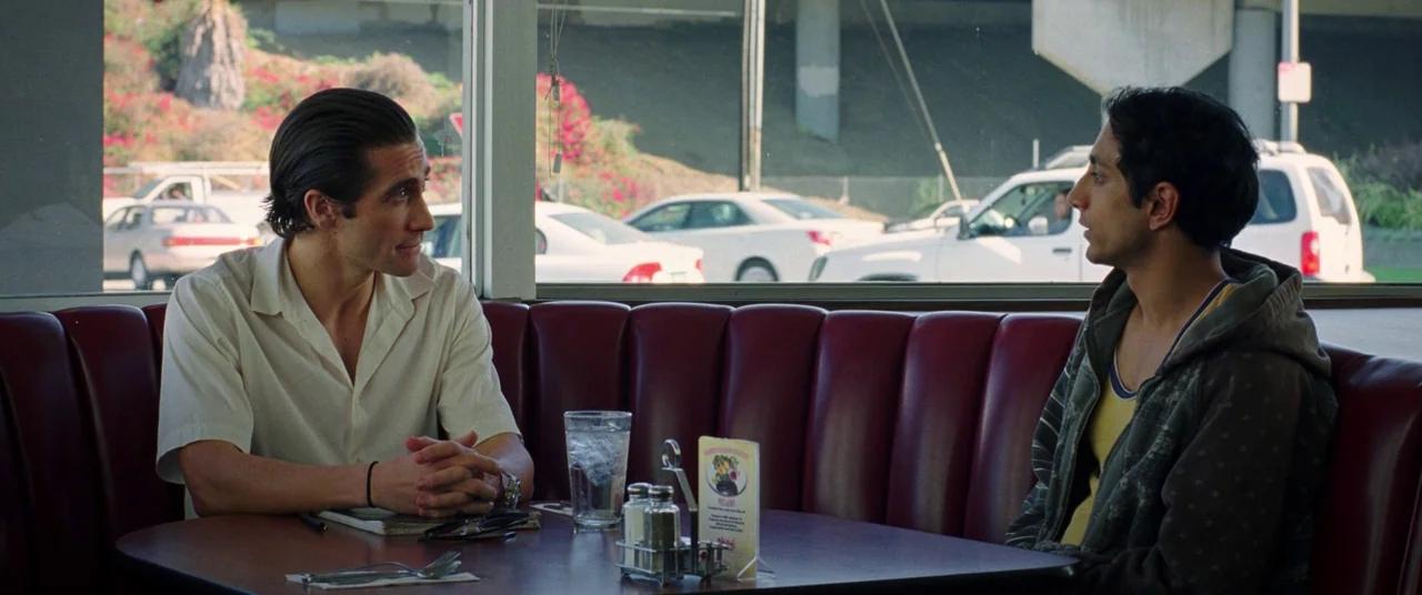 Nightcrawler (2014) movie still: medium — A classic diner booth with red vinyl seating positioned near a large glass window. Two men sit opposite…