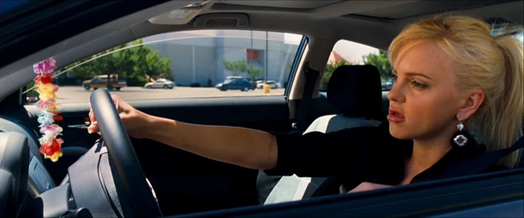 Observe And Report (2009) movie still: medium — A woman drives a car through a parking lot with a large building visible. She keeps both hands on…