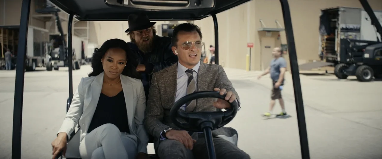 Last Looks (2022) movie still: medium — A man drives a golf cart with two passengers in a paved industrial area. The background features…