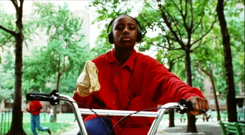 Clockers (1995) movie still: medium — An urban park setting features leafy deciduous trees and scattered green benches. A teenager rides a bicycle…
