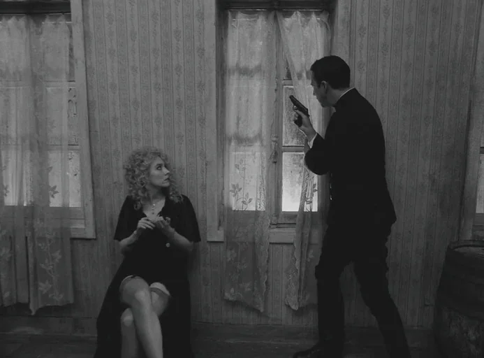 The French Dispatch (2021) movie still: medium — A room with patterned wallpaper features tall windows covered by thin lace curtains. A man points a…