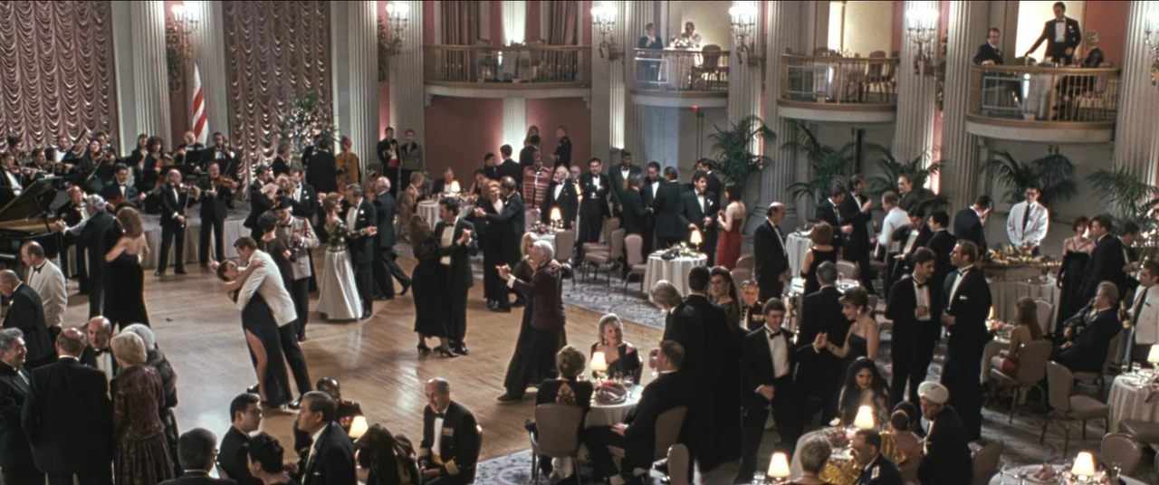 True Lies (1994) movie still: wide — A large, elegant ballroom filled with numerous guests in formal evening wear during a gala. Musicians play on a…