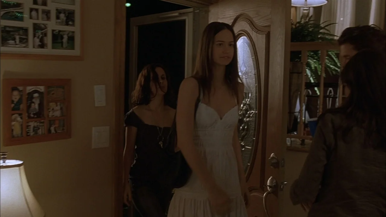 The Babysitters (2008) movie still: medium — A dimly lit home entryway features a wooden door and a picture-filled wall. Two young women stand in…