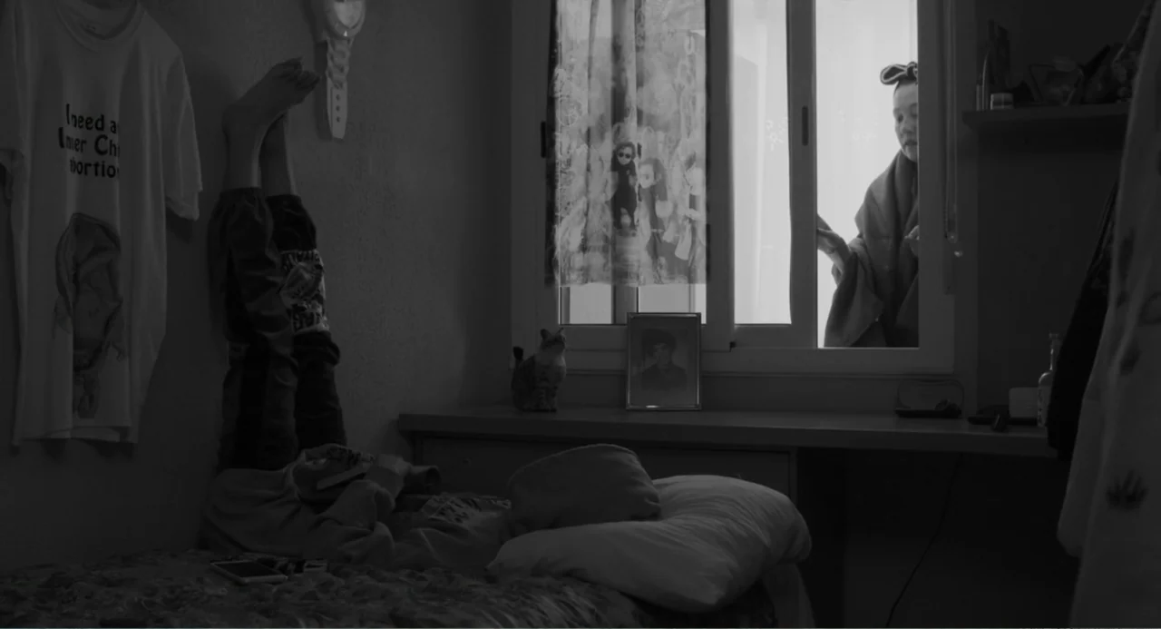 El Planeta (2021) movie still: medium — A dimly lit room contains a person resting on a bed with feet elevated on the wall. A second person stands…