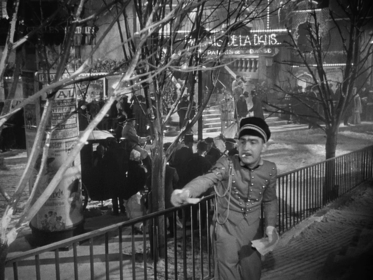 Le Plaisir (1952) movie still: medium — A busy night street outside a dance hall with bare trees and posters. A man in a uniform stands near a metal…