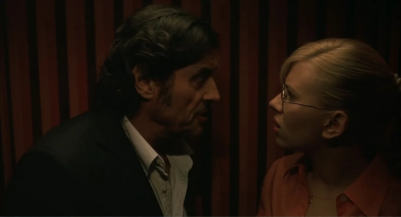 Scoop (2024) movie still: medium — A confined elevator interior features dark vertical wood-paneled walls and low ambient lighting. An adult man and…