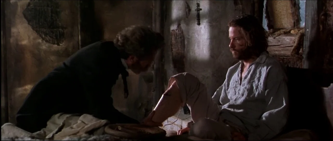 Ravenous (1999) movie still: medium — A dimly lit room with distressed plaster walls and rough textures. One man tends to the leg injury of another…