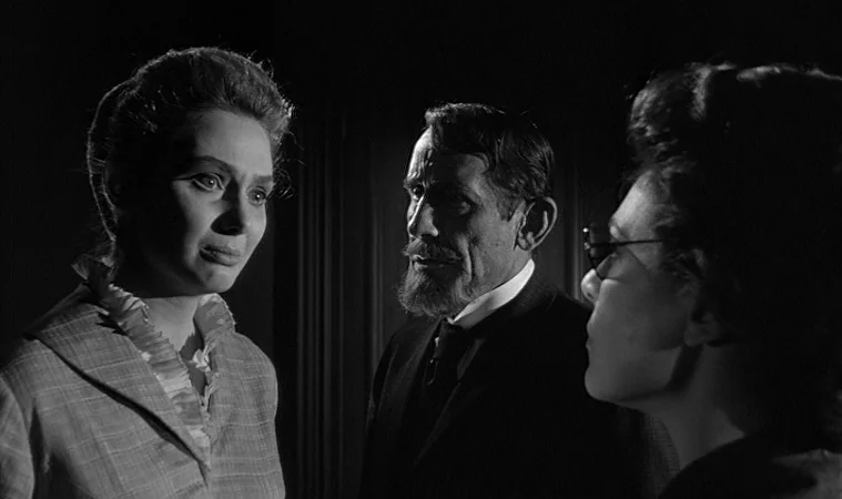 The Miracle Worker (1962) movie still: medium — A dark, dimly lit indoor environment with heavy shadows obscuring the background wall. Three people…