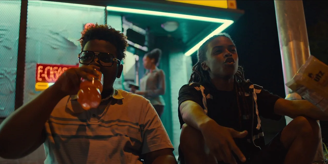 Charm City Kings (2020) movie still: medium — Two teenage boys sit outside a convenience store illuminated by bright cyan neon lights. One boy…