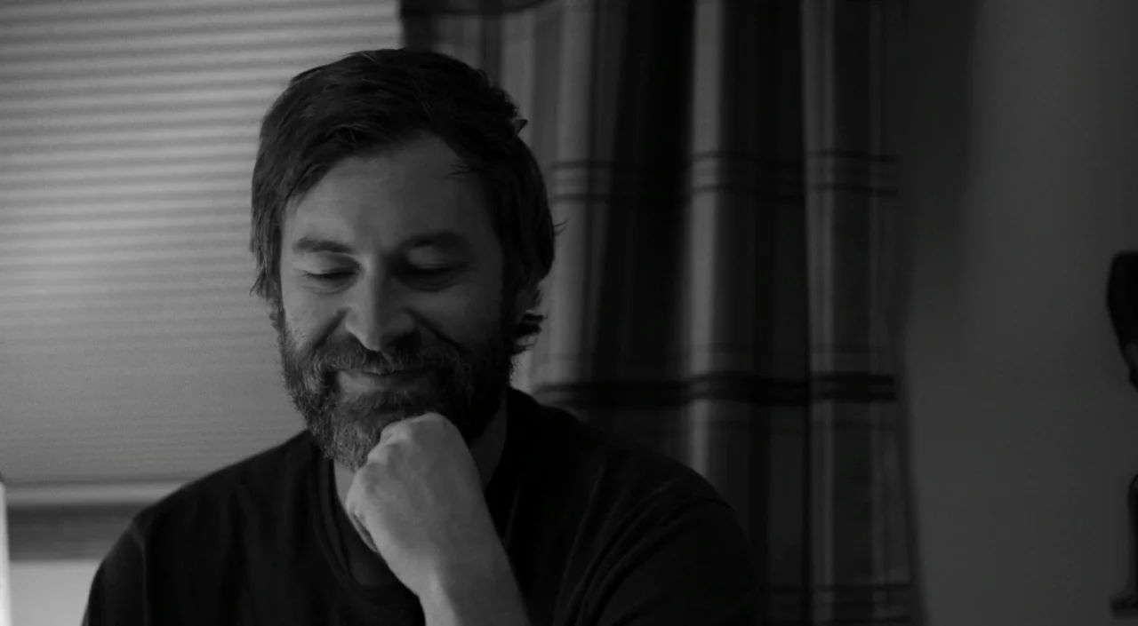 Blue Jay (2016) movie still: closeup — A residential room interior with horizontal blinds and fabric curtains partially visible. A man rests his…