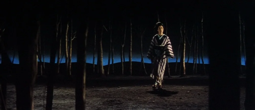 An Actor’s Revenge (1963) movie still: fullbody — A dark clearing features sparse, thin trees silhouetted against a sharp blue horizon. A woman…