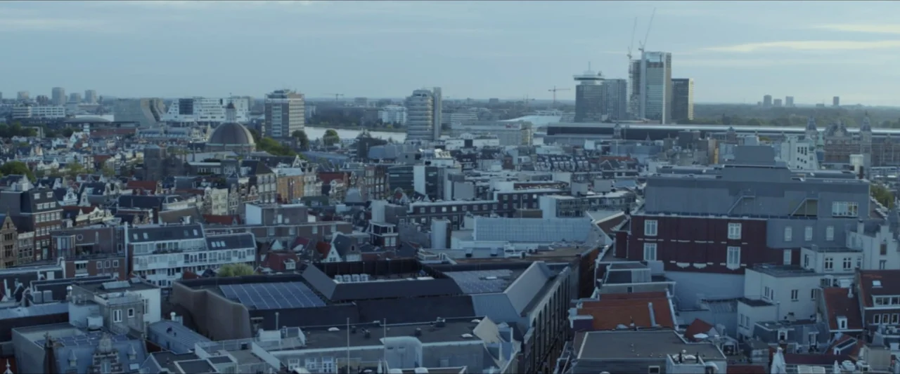 Black Lotus (2023) movie still: establishing — A dense urban expanse of mixed architectural styles extends toward the distant horizon line. Numerous…