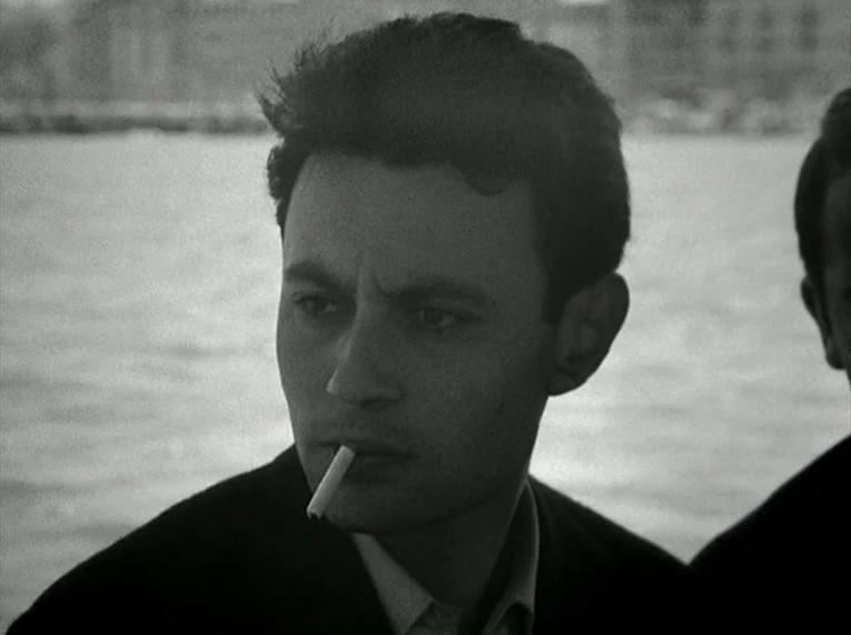 Le Petit Soldat (1963) movie still: closeup — A man stands outdoors near a body of water with buildings visible in the distance. He stares into the…