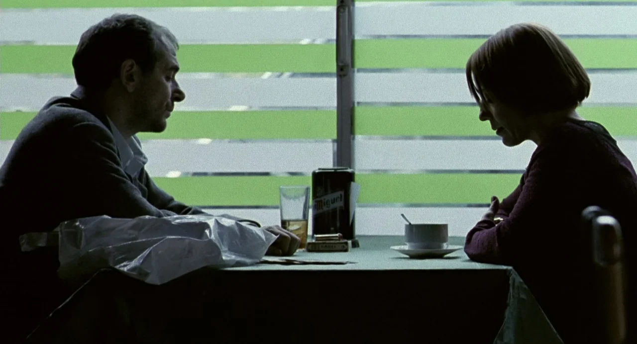 The Nameless (1999) movie still: medium — A table set against a large window with green-tinted privacy slats and metallic framing. A man and a woman…