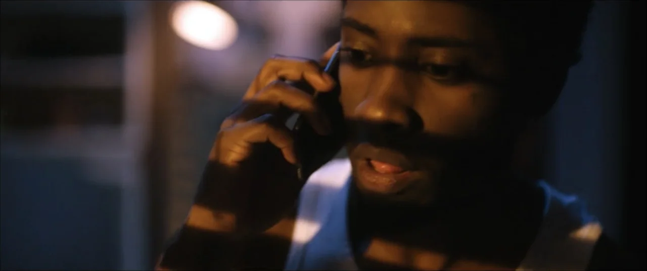 I'll Take Your Dead (2018) movie still: closeup — A dimly lit interior space with shadows cast by window blinds across the wall. A young man in a…