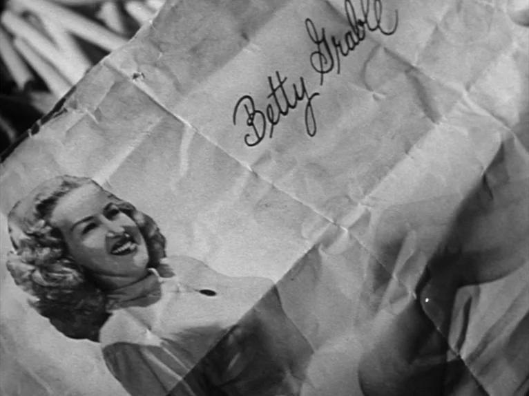 Stalag 17 (1953) movie still: closeup — A crinkled piece of paper features a printed black and white image of Betty Grable. The handwritten…