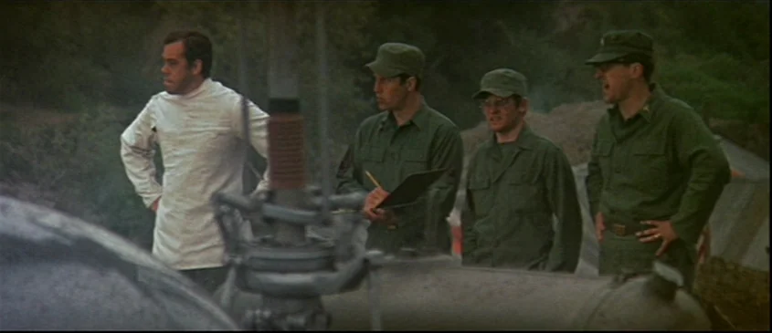 MASH (1970) movie still: medium — An outdoor military camp environment with tents visible in the blurry background. Four men stand together near…