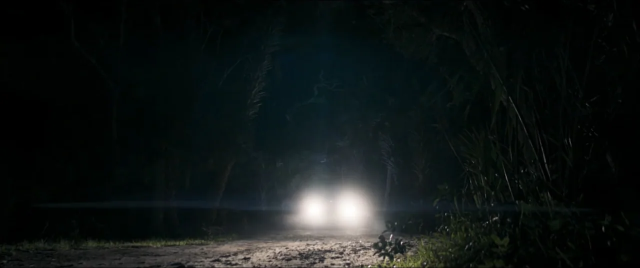 Offseason (2022) movie still: wide — A narrow dirt track winds through a dense, dark forest at night. Two bright vehicle headlights pierce the deep…