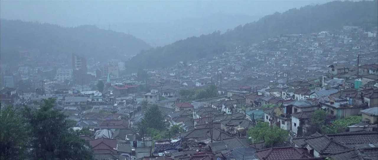 Sympathy for Mr. Vengeance (2002) movie still: establishing — A dense hillside neighborhood features traditional dark tiled roofs under a heavy rain…