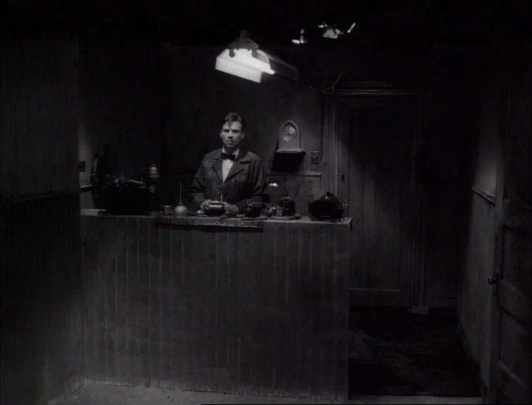 Eraserhead (1977) movie still: medium — A dimly lit industrial space features a wooden workbench against a dark wall. A man in a work jacket stands…