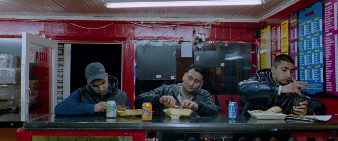Catch Me Daddy (2014) movie still: medium — A red-walled takeaway shop interior features a black counter and a printed menu board. Three men sit in…