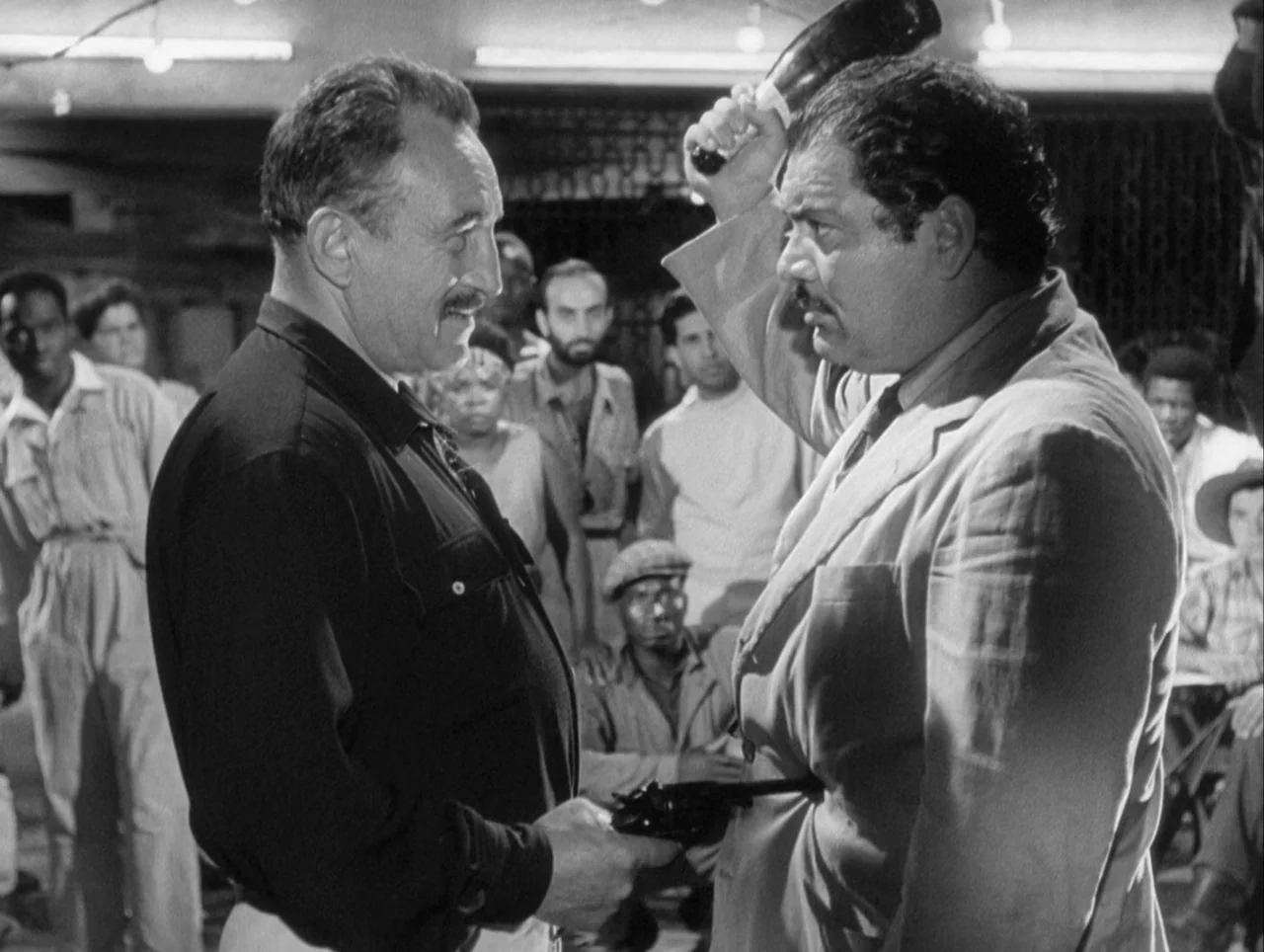 The Wages of Fear (1953) movie still: medium — A crowded interior space with men in work clothes gathered in the background. One man holds a…
