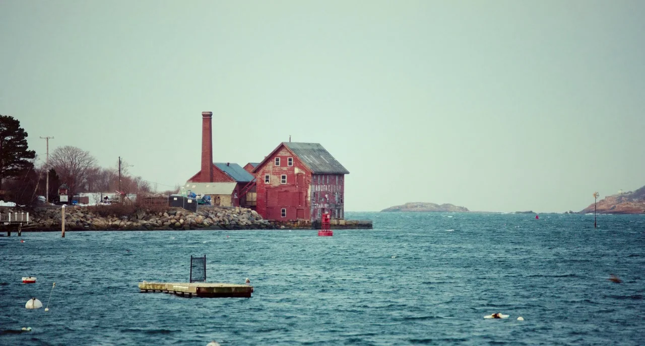 Manchester By The Sea (2016) movie still: establishing — A weathered red brick factory with a smokestack stands on a rocky coastal edge. The…