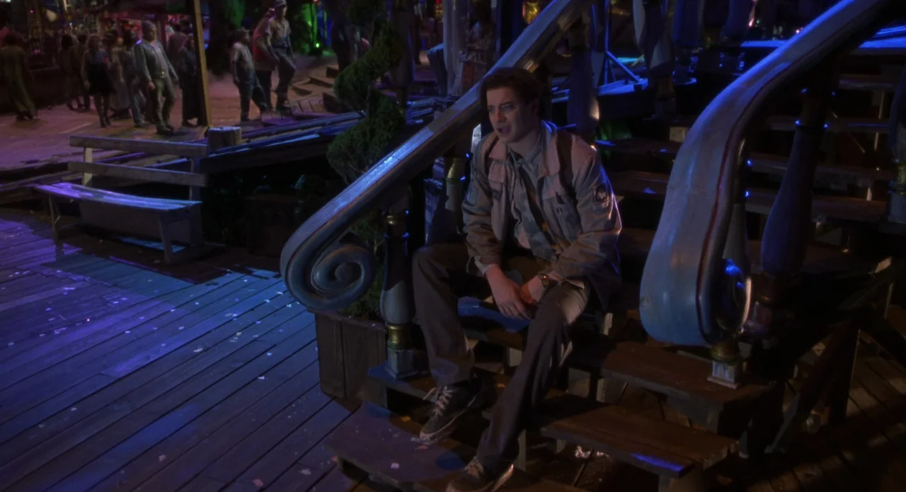 Monkeybone (2001) movie still: medium — A man sits on a dark wooden staircase at night in a populated outdoor area. The surrounding environment is…