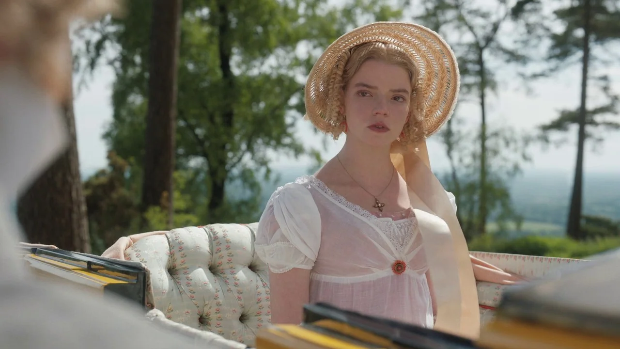 Emma. (2020) movie still: medium — A woman in 19th-century attire sits in an open carriage with tufted upholstery. She looks towards another person…