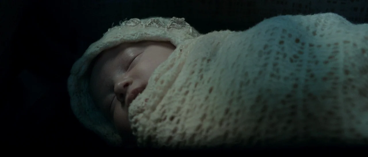 Predestination (2014) movie still: closeup — A dark space focuses entirely on an infant resting in a soft swaddle. The baby sleeps peacefully,…