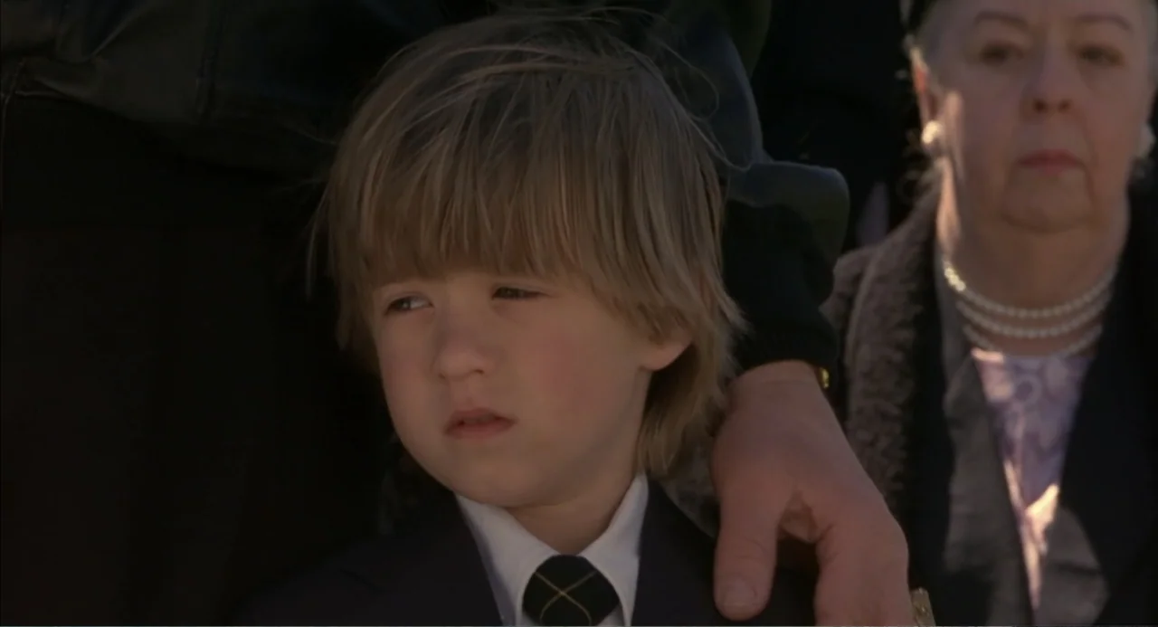 Bogus (1996) movie still: closeup — A group of people dressed in formal dark attire stand together outdoors. A young boy in a blazer looks forward…