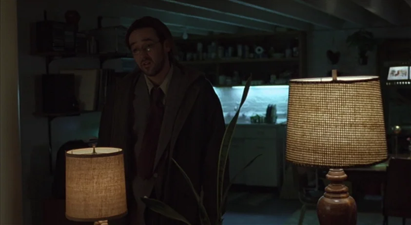 Being John Malkovich (1999) movie still: medium — An interior space with dim, low-key lighting focused on two lamps in the foreground. A man stands…
