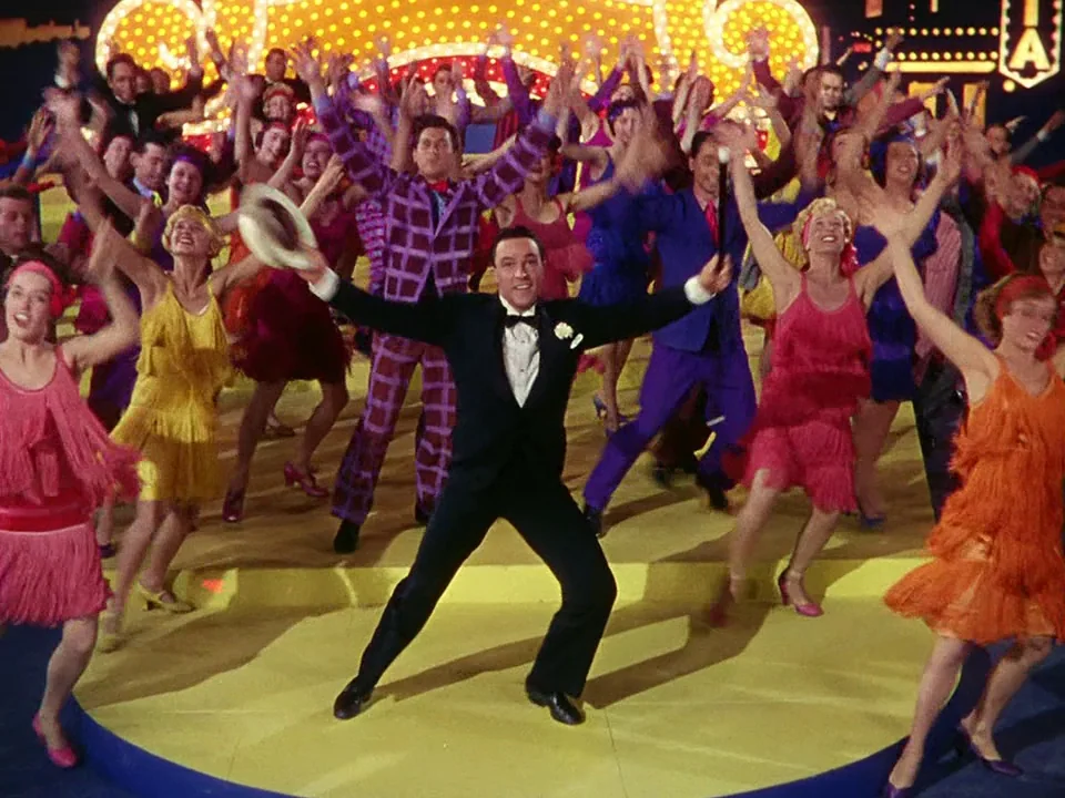 Singin’ in the Rain (1952) movie still: fullbody — A brightly lit theatrical stage features a group of dancers in colorful period costumes. A man in…