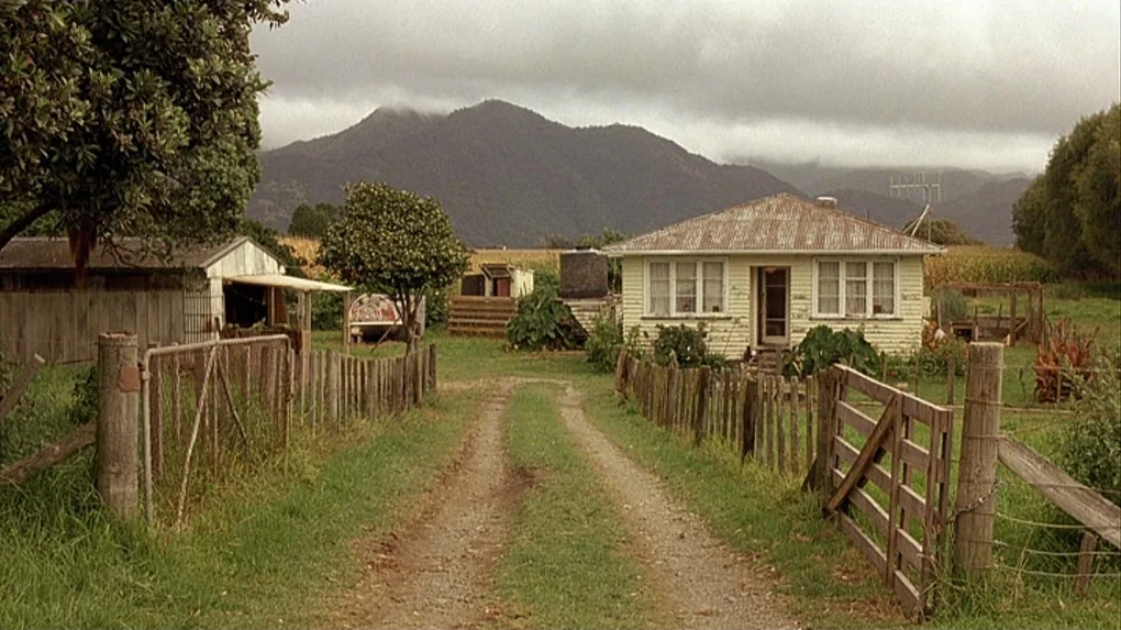 Boy (2010) movie still: wide — A rural farm with a gravel path leading toward a small, light-colored wooden house. Cloudy mountains rise in the…