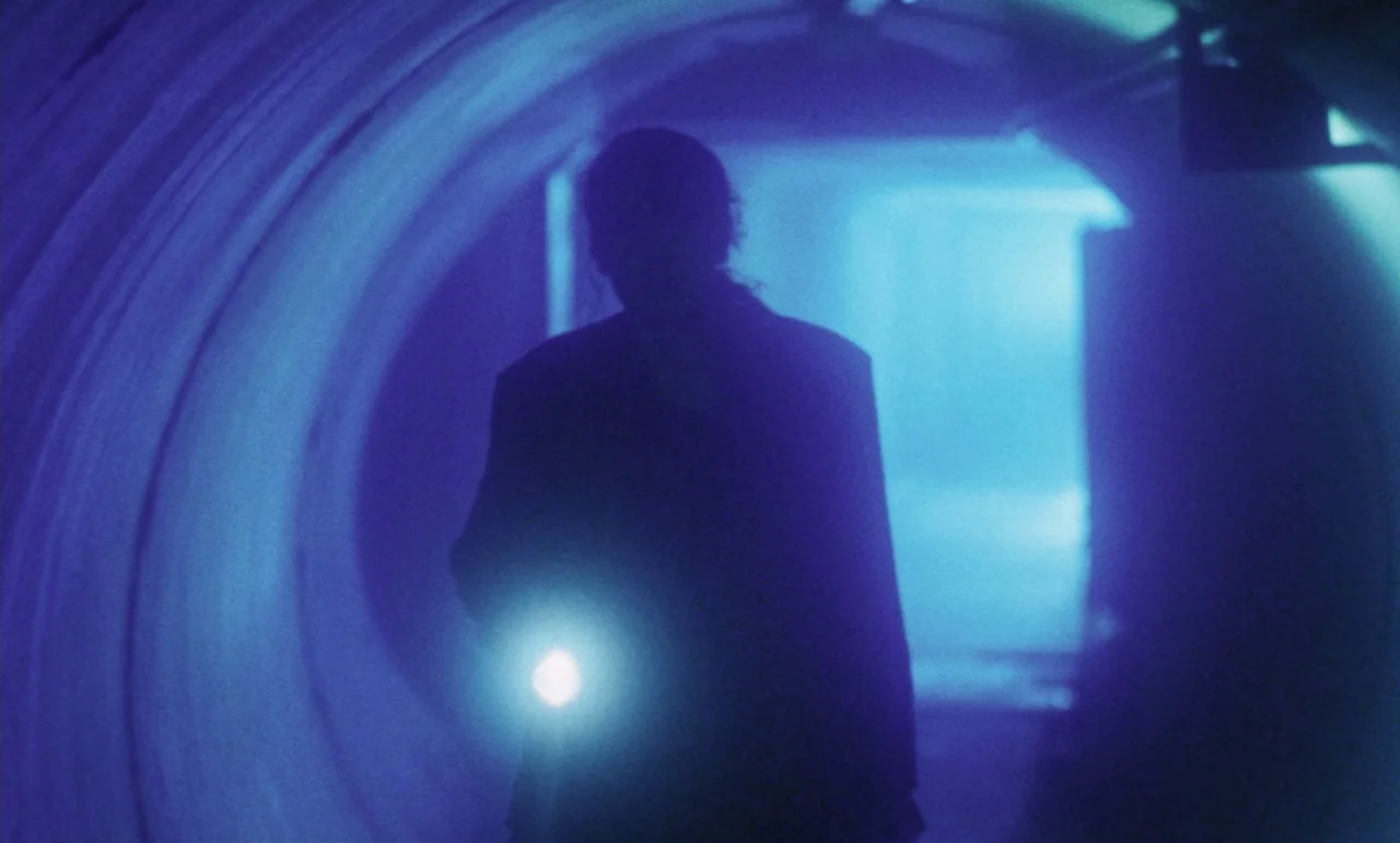 Evil Dead Trap (1988) movie still: medium — A person walks through a curved, concrete tunnel illuminated only by their handheld beam. The silhouette…