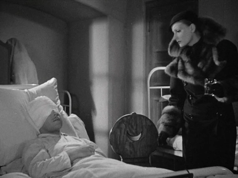 Mata Hari (1931) movie still: medium — A dimly lit hospital room features a metal bed frame and simple wooden chair. A woman in a fur-trimmed coat…