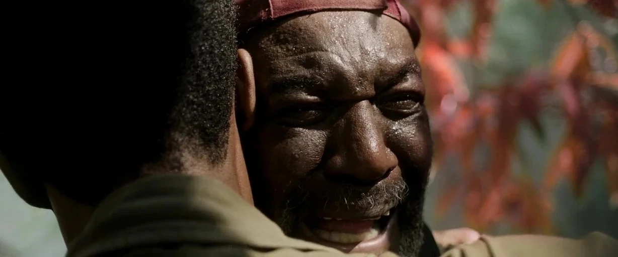 Da 5 Bloods (2020) movie still: closeup — An outdoor setting with foliage visible in the blurred background. An older man with a grey-flecked beard…