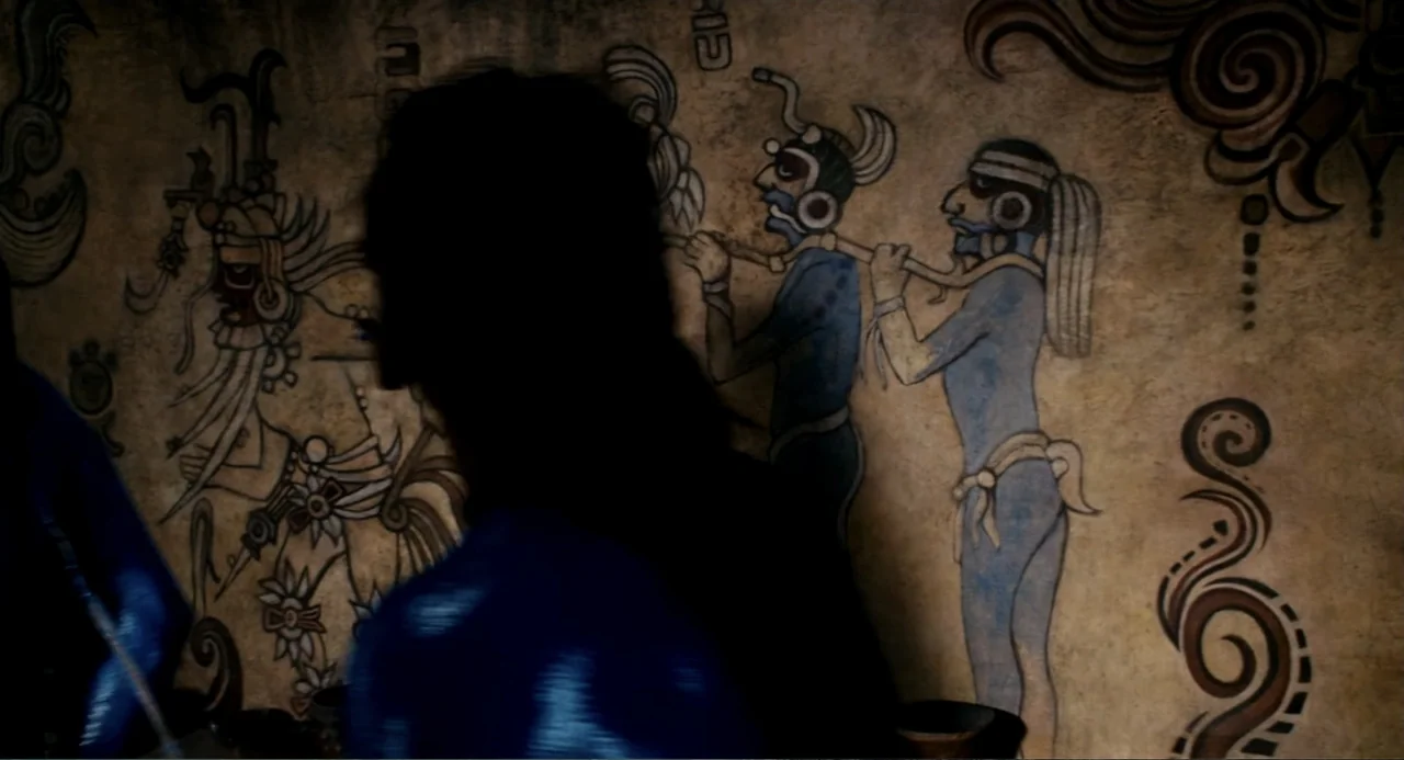 Apocalypto (2006) movie still: medium — A dark stone wall features a painted mural of ancient figures in blue pigment. An obscured person in the…