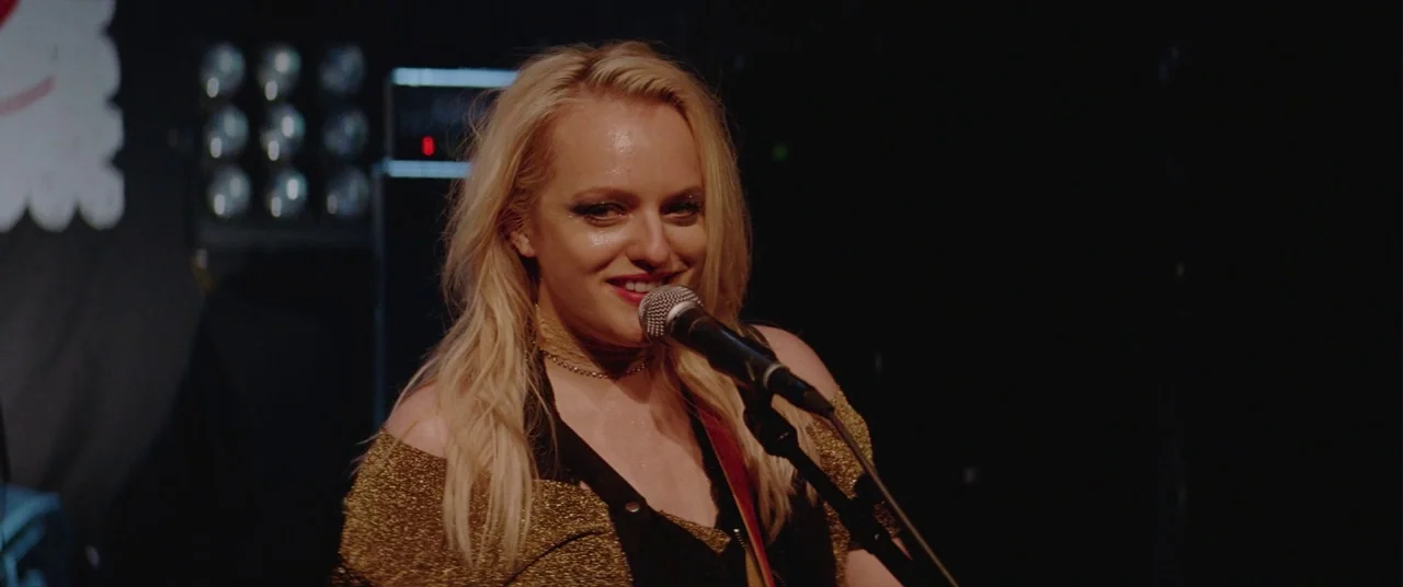Her Smell (2018) movie still: medium — A woman stands on a dark stage with blurred lights visible in the background. She is singing into a…