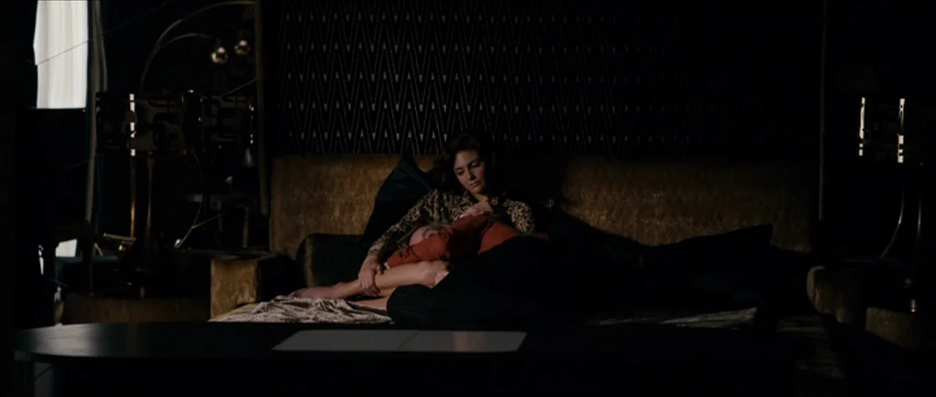 The Look of Love (2013) movie still: medium — A dark bedroom with patterned wallpaper and a large cushioned headboard. A woman sits on the bed while…